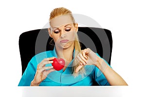 Worried female doctor or nurse sitting behind the desk holding heart model and showing thumb down