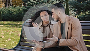 Worried hispanic young couple with laptop sit on bench in autumn park unsuccessful online shopping broken device serious