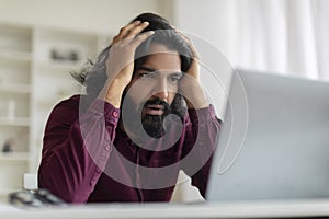 Worried indian man clutching head in frustration while staring at computer screen