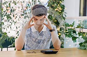 Worried male employee holding his hand behind his head and looking at the desk where the calculator and money lie while sitting at