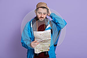 Worried man lost hiking, looking at map with confused facial expression, wearing jacket and hat, keeping hand on head, posing