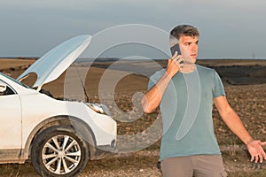 A worried man in the middle of the field calling the insurance of his broken down car for assistance at sunset