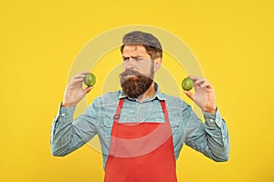 Worried man in red apron looking at fresh limes citrus fruits yellow background, fruiterer