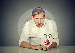 Worried man with sad expression holding clown mask expressing cheerfulness