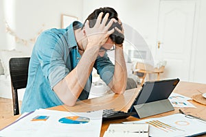 Worried man sitting at the table at home, holding his head, worried about his bills. Loss of money in investments.