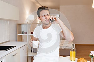 Worried man in white t-shirt talking on phone holding cup of tea in kitchen. Serious brunette guy standing beside table