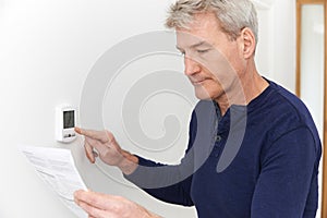 Worried Mature Man With Bill Turning Down Heating Thermostat