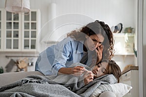 Worried mother looking at thermometer checking temperature of sick child daughter lying in bed