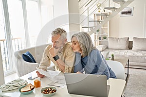 Worried old senior couple checking bank documents or bills at home.
