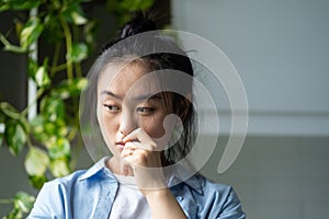 Worried pensive Asian woman touching lips nervous stands in apartment near closet with houseplants