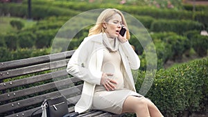 Worried pregnant female calling 911 feeling pain in belly, sitting on park bench
