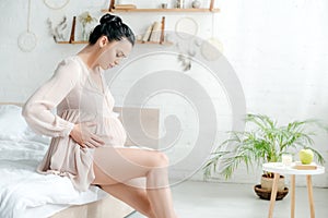Worried pregnant woman in nightie feeling pain and touching belly while sitting on bed