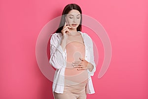 Worried pregnant woman talking on phone with her doctor, being concentrated, keeps hand on her belly, standing isolated over rosy