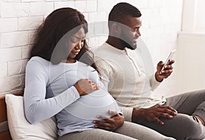 Worried pregnant woman touching tummy while indifferent husband using phone