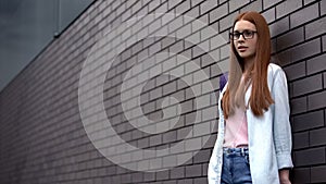 Worried red-haired female teenager standing campus wall, university difficulties