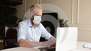 Worried redhead bearded man wearing protective medical mask working with a laptop