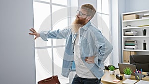 Worried redhead young man, a hard-working office worker, suffering terrible backache while standing in his indoor workplace