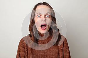 Worried scared brown haired adult woman wearing brown sweater posing isolated over gray background looking at camera with big eyes