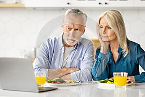 Worried senior couple reading news on laptop during breakfast in kitchen