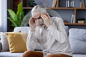 Worried senior gray-haired man suffering from headache at home on sofa, holding hands to head