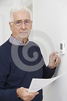 Worried Senior Man With Bill Turning Down Central Heating Thermostat