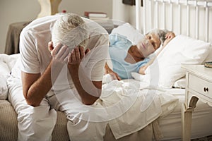 Worried Senior Man Sits On Bed Whilst Wife Sleeps