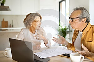 Worried senior spouses checking financial documents with laptop, sitting in kitchen, suffering problems