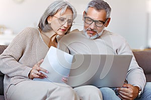 Worried serious senior family couple paying bills online on laptop while sitting on sofa at home