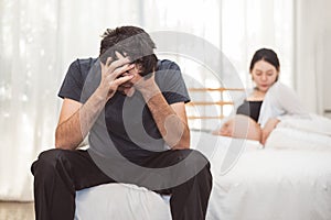 Worried stress man sitting on bed with hand on forehead in bedroom in serious mood emotion with pregnant wife woman background.