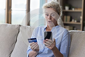 Worried surprised elderly woman holding card, look at smartphone screen
