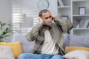 Worried and upset young hispanic man scared by loud noise and scream sitting on sofa at home and covered ears with hands