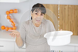 Worried woman holding bucket while water droplets leak from ceiling