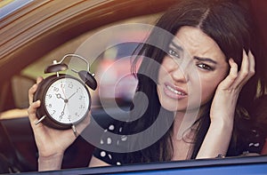 Worried woman inside car showing alarm clock running late to work