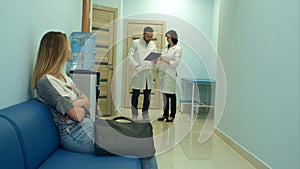 Worried woman patient waiting in hospital hall while two doctors discussing diagnosis