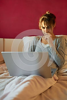 Worried woman sitting on bed at home, frowning and watching laptop computer, browsing on Internet.