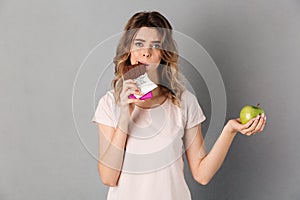 Worried woman in t-shirt eating chocolate while holding fresh apple