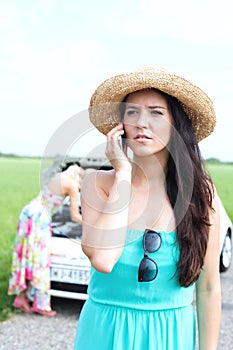Worried woman using mobile phone while friend examining broken down car at countryside