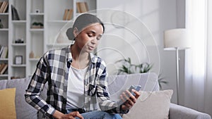 Worried young African American girl sitting on the sofa looking at her phone and not understanding why it is not working