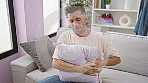 Worried young hispanic man, portrait of illness, suffering intense stomachache while unhappily resting, lying on living room sofa