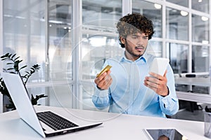 Worried young hispanic man sitting at the desk in the office and looking at the phone upset, holding a credit card in