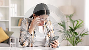 Worried young Indian girl sitting at the table looking at her phone and does not understand why it is not working. The