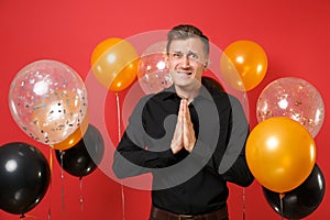 Worried young man in black classic shirt folded his hands and praying on red background air balloons. St. Valentine`s