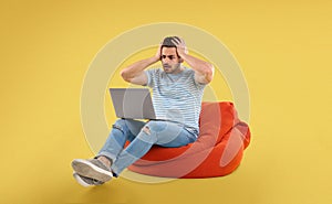 Worried young man with laptop on bean bag chair, yellow background