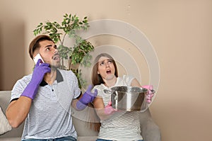 Worried young woman collecting water leakage from ceiling while her husband calling plumber