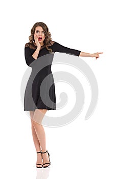 Worried Young Woman In Elegant Black Dress And High Heels Is Pointing At The Side