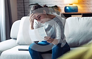 Worried young woman looking at bills and thinking about her problems while sitting on the sofa at home
