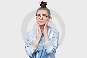 Worried young woman, looking directly at the camera through round eyewear. Doubtful girl wears blue shirt can`t make choice, has