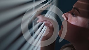 Worried young woman looking through window blinds into darkness. Paranoia concept