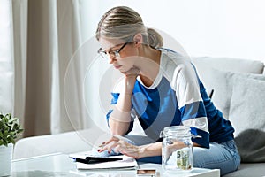 Worried young woman using calculator and counting her savings while sitting on sofa at home