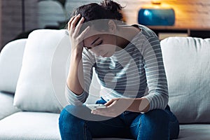Worried young woman using her mobile phone while sitting on sofa in the living room at home